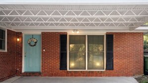 Una casa de ladrillo con una puerta verde azulada, dos ventanas con contraventanas y un porche cubierto.