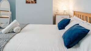 Een net opgemaakt bed met wit en blauw beddengoed, een klein nachtkastje met een lamp, en een spiegel aan de muur.