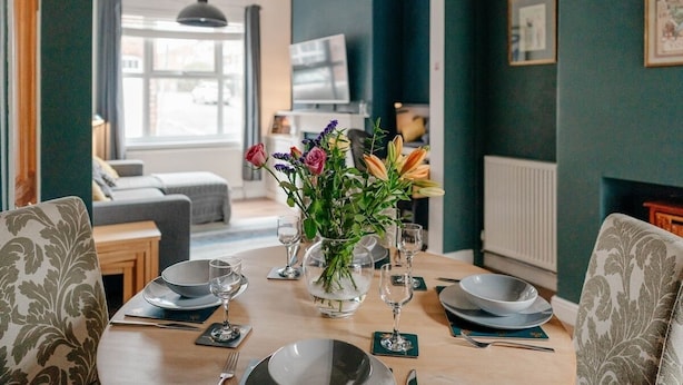 Een eethoek met een tafel gedekt voor vier personen, een bloevas, en zicht op een woonkamer en keuken op de achtergrond.