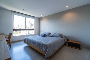 Ein modernes Schlafzimmer mit einem großen Bett, einem Nachttisch aus Holz und Blick auf eine Stadtlandschaft durch ein großes Fenster.