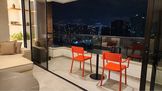 A balcony with a city view at night, featuring a sofa, two orange chairs, and a table.
