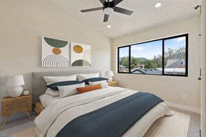 Une chambre à coucher comprenant un lit, des tables de chevet, un ventilateur de plafond, des œuvres d’art et une fenêtre donnant sur des maisons et des arbres.