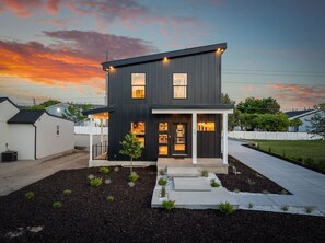 Une maison moderne à deux étages, avec une façade sombre, un porche à l’avant et un jardin bien entretenu.