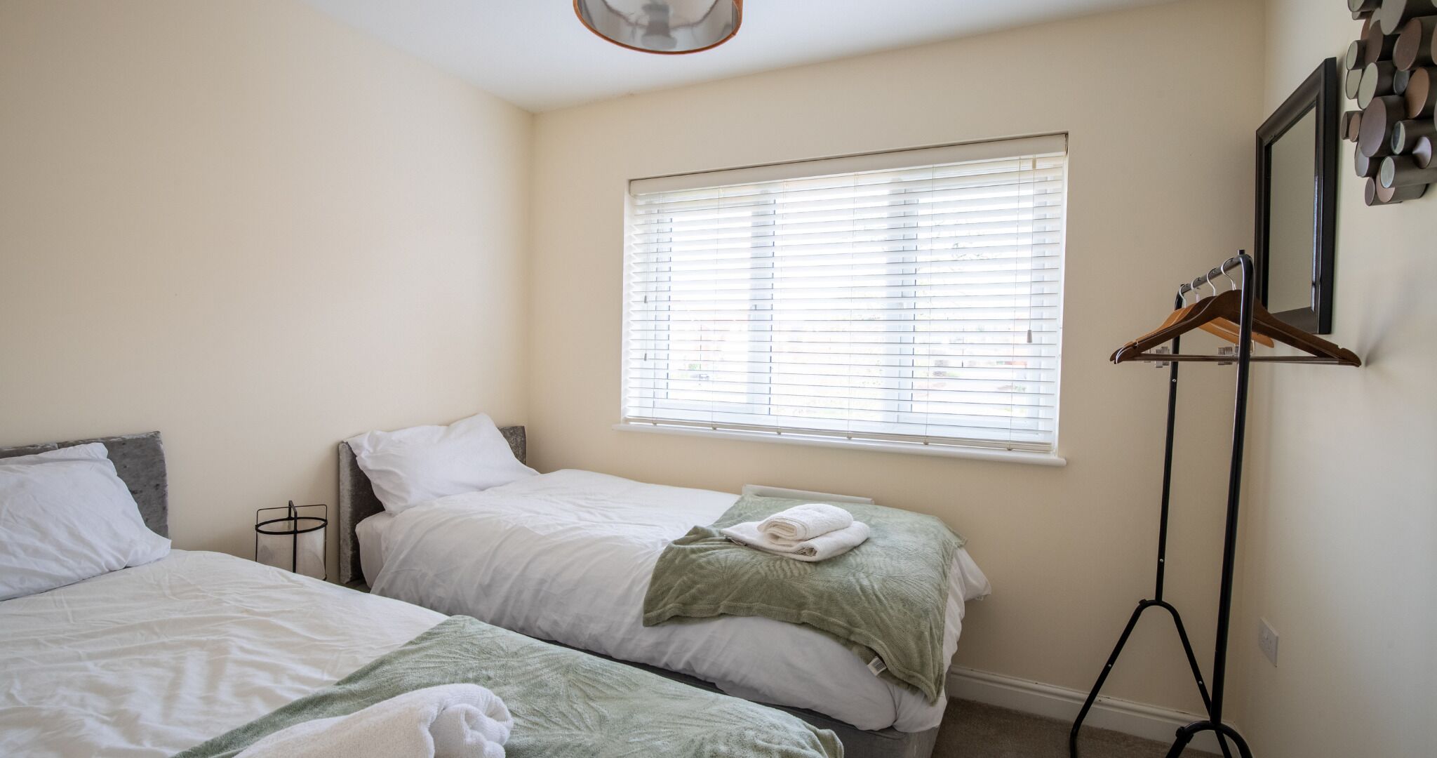 Een slaapkamer met twee bedden, een raam met zonwering, een kapstok en een spiegel aan de muur.