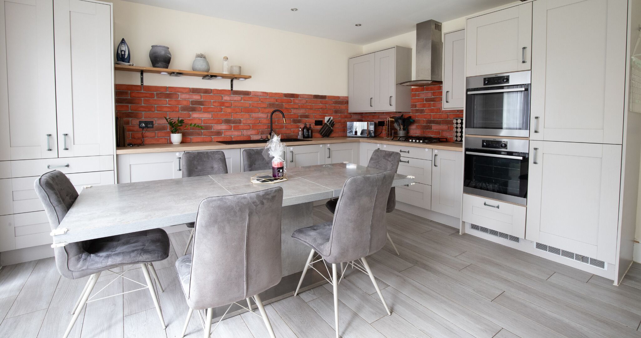 A modern kitchen with a brick backsplash, white cabinetry, and a dining area with grey upholstered chairs.