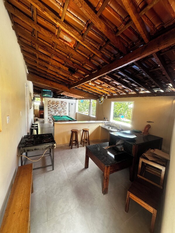 A rustic kitchen with a wooden ceiling, a gas stove, stools, a black countertop, and a window with a view of greenery.