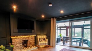 A living room with a stone fireplace, a comfortable armchair, a dining table, and a large window offering a view of the outdoors. - 7 Brewery Pool (Newton Stewart)
