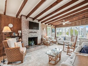 A cozy living room with a fireplace, wicker furniture, and a large window overlooking a residential area. - Spacious Bethany Beach Home w/ Deck (Bethany Beach)