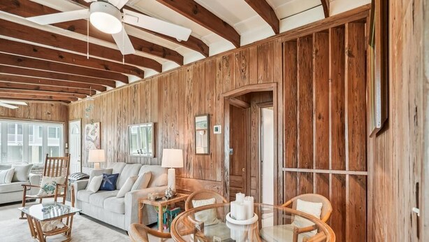 Ein rustikales Interieur mit freiliegenden Holzbalken, einem runden Glastisch, Korbstühlen, einer Couch und einem großen Fenster.