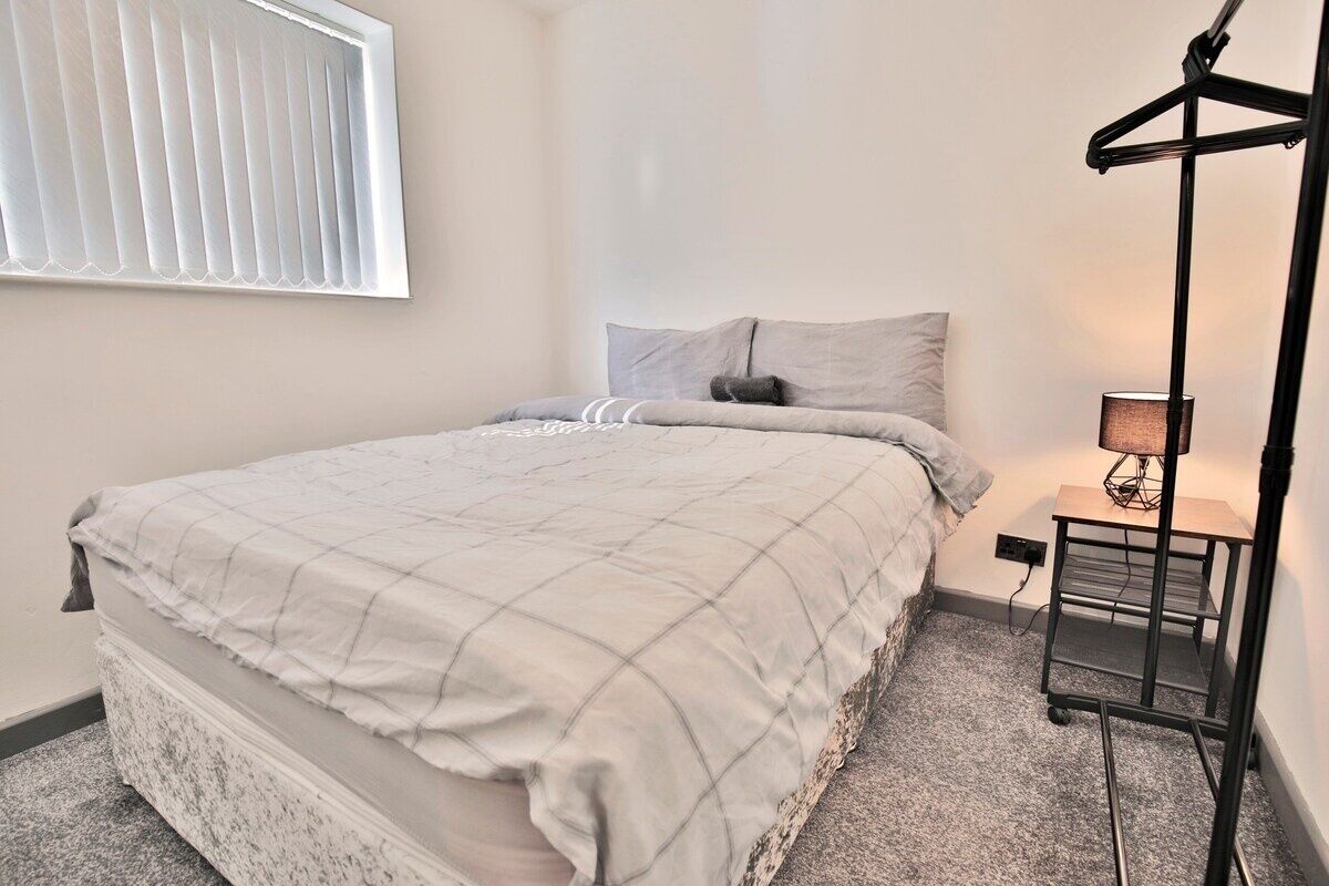 A bedroom with a bed, a bedside table with a lamp, a window with blinds, and a wall-mounted radiator.