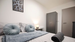 Un lit bien fait, avec une couverture grise et deux oreillers assortis, une lampe de chevet, une armoire grise et une porte sur la droite.