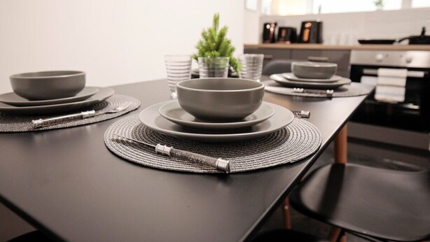 Une cuisine moderne avec une table à manger dressée pour quatre personnes, agrémentée d’assiettes et de bols gris, d’une petite plante en pot et d’un plan de travail en arrière-plan.