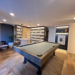 A modern pool table in a room with a built-in shelving unit, a small seating area, and a potted plant.