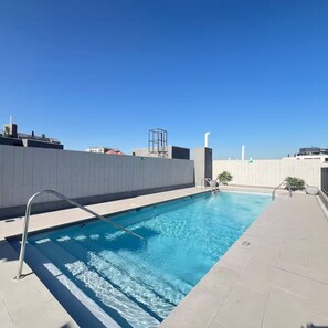 A rooftop swimming pool with clear blue water, surrounded by a white tiled deck and a grey wall.