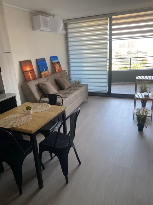 A modern living room with a sofa, a dining table with chairs, and a large window with blinds.