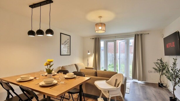 A dining area with a wooden table set for four, a sofa, a white high chair, and a television displaying 'NETFLIX'. - Littlebrooke House | Bolton | Free Parking (Bolton)