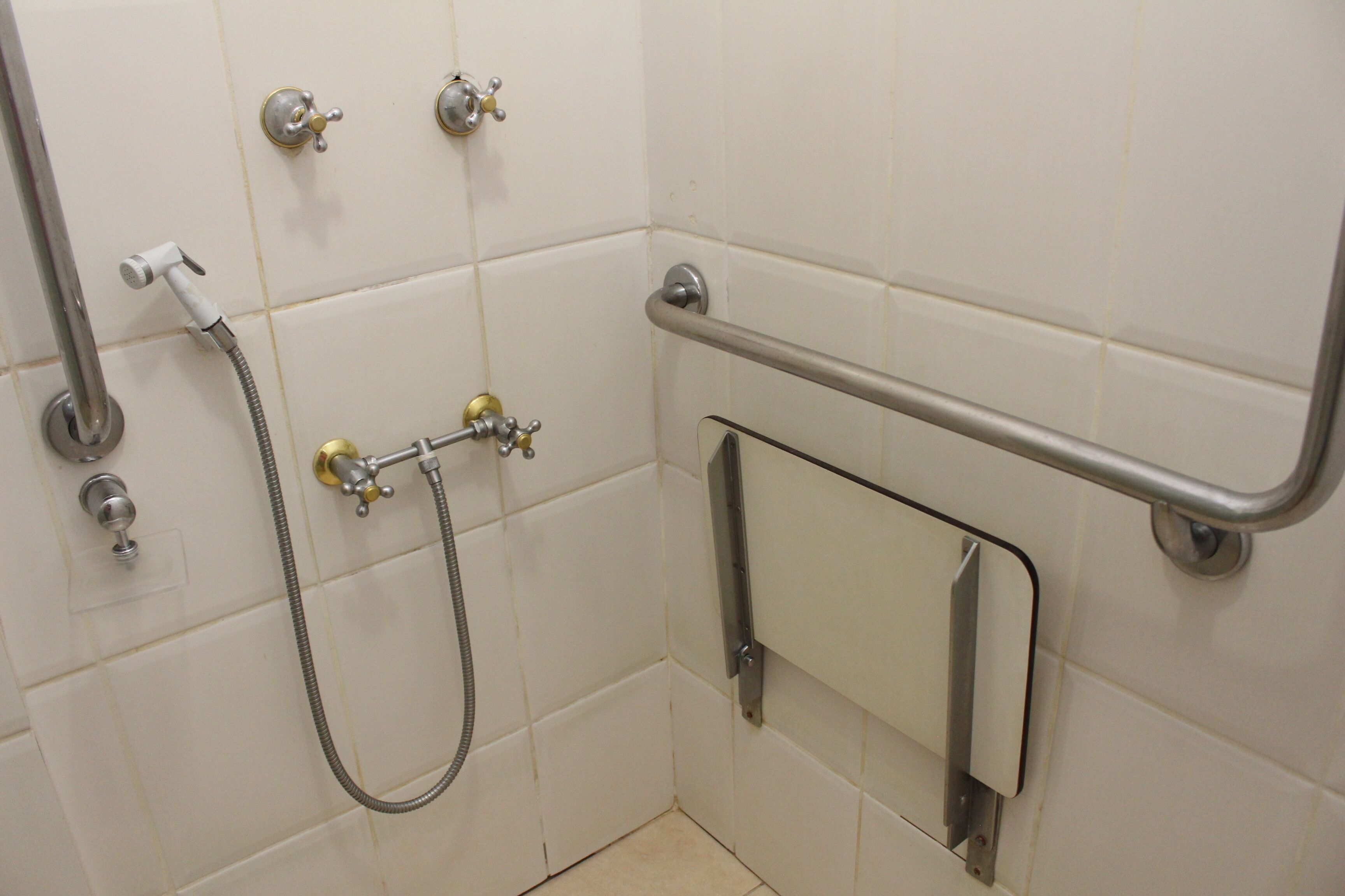 Casa de banho com azulejos brancos, chuveiro com chuveiro manual, barra de apoio e espelho.
