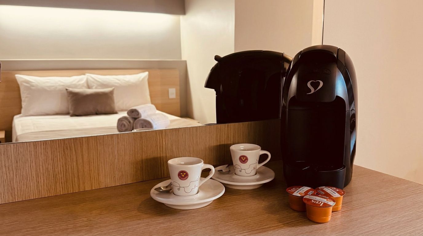 Habitación de hotel con cama, cafetera, dos tazas y un espejo que refleja la habitación.