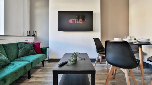 Ein modernes Wohnzimmer mit einem grünen Sofa, einem schwarzen Couchtisch und einem Essbereich mit Tisch und Stühlen. Ein an der Wand montierter Fernseher zeigt das Netflix-Logo.