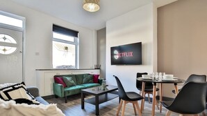 Ein modernes Wohnzimmer mit einem grünen Sofa, einem Couchtisch und einem Essbereich mit Stühlen und Tisch. Ein an der Wand montierter Fernseher zeigt das Netflix-Logo.