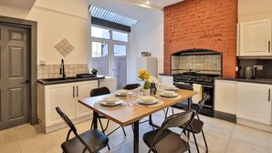 A modern kitchen with white cabinets, a brick accent wall, a dining table set for four, and a large window providing natural light. - Chesterfield House, 7BR, Blackpool (Blackpool)