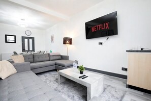 Una sala de estar moderna con un sofá seccional gris, una mesa de centro, un televisor en la pared mostrando 'NETFLIX', una lámpara y una puerta.
