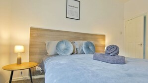 Una cama bien hecha con una colcha azul claro, dos almohadones decorativos, una toalla enrollada y una mesita de noche con una lámpara.