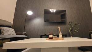 Ein modernes Wohnzimmer mit einem an einer dunklen Wand montierten Flachbildfernseher, einem weißen Couchtisch mit Kerze und kleiner Pflanze sowie zwei schwarzen Ledersofas.