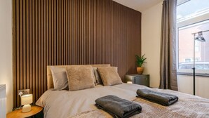 A neatly made bed with a textured blanket and two folded towels, a bedside lamp, a small table with a lamp, and a potted plant on a nightstand.