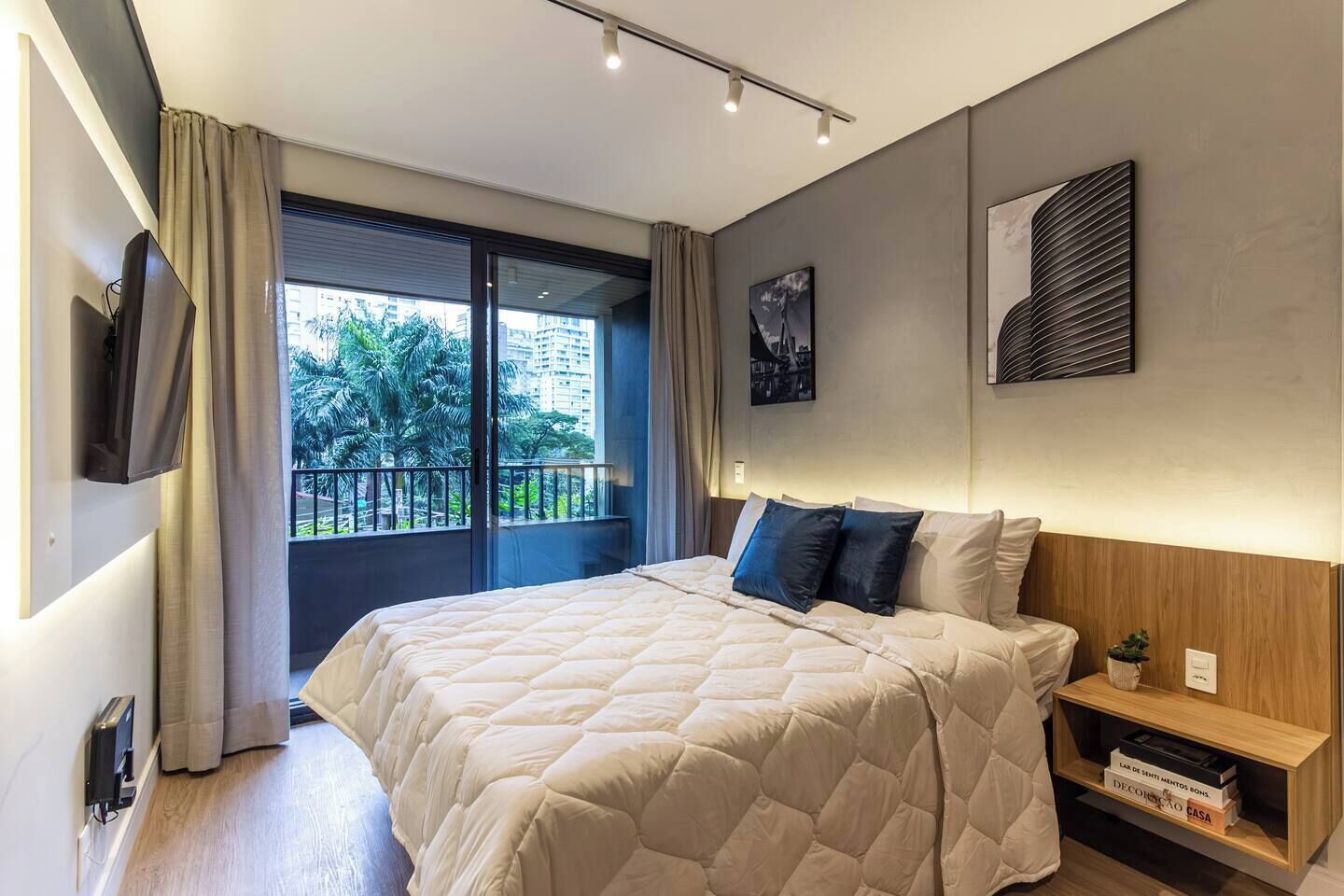 Een moderne hotelkamer met een groot bed, een aan de muur bevestigde televisie, een balkon met uitzicht op palmbomen en een boekenkast met boeken.