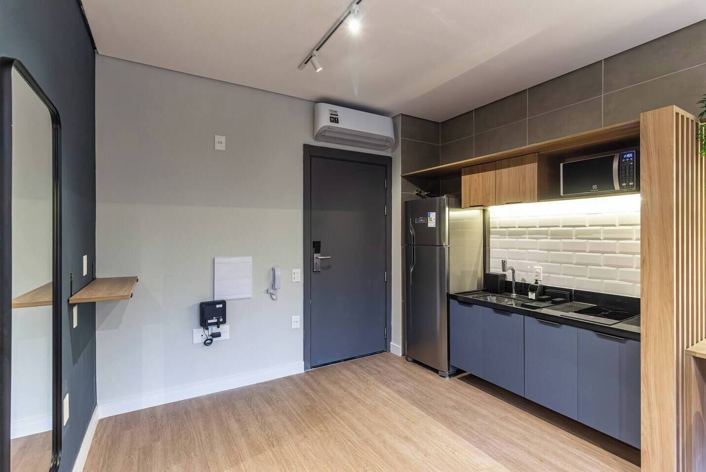 A modern kitchen with grey cabinets, a stainless steel refrigerator, and a microwave. There is a wooden shelf above the sink and a small window above the door.