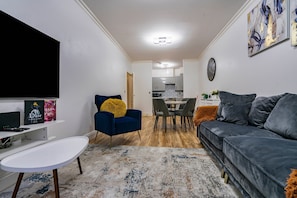 A modern living room with a grey sofa, a blue armchair, a white coffee table, and a dining area with a kitchen in the background. - Old Market House, Manchester, Sleeps 6 (Manchester)