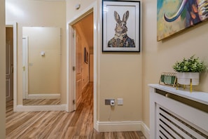 A hallway with a wooden floor, a framed artwork of a rabbit, a potted plant, and a calendar on a shelf. - Old Market House, Manchester, Sleeps 6 (Manchester)