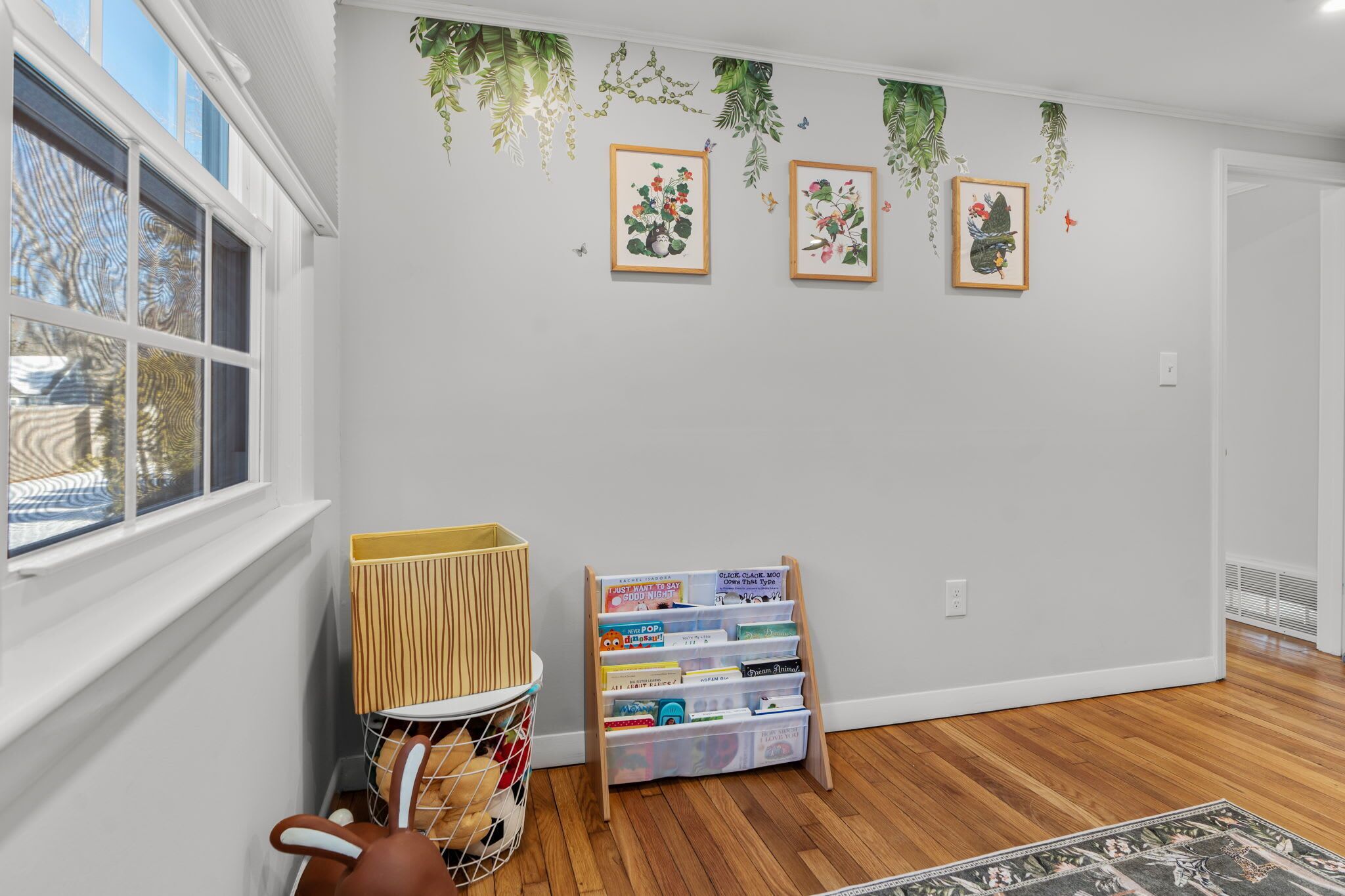 Una habitación con piso de madera, paredes blancas, una ventana con persianas y un estante en la pared con fotografías enmarcadas.
