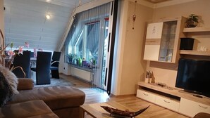 A modern living room with a sofa, a coffee table, and a TV. There is a window with curtains, a plant on a shelf, and a staircase leading to another room. - NEW! Modern living in Erbendorf - Upper Palatinate (Erbendorf)