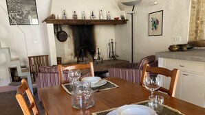 Une salle à manger avec une table en bois dressée pour quatre personnes, entourée de chaises à rayures. On peut voir une cheminée avec un foyer en métal et une étagère au-dessus, où sont disposées diverses bouteilles et objets décoratifs.