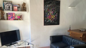 Un salon avec un téléviseur posé sur une table en verre, un canapé en cuir noir, un bureau en bois et un mur orné d’un tableau noir et de livres.