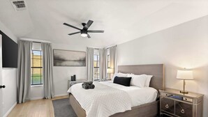 Ein Schlafzimmer mit einem Bett, einem Nachttisch, einer Lampe, einem Deckenventilator und einem Fenster mit Vorhängen.