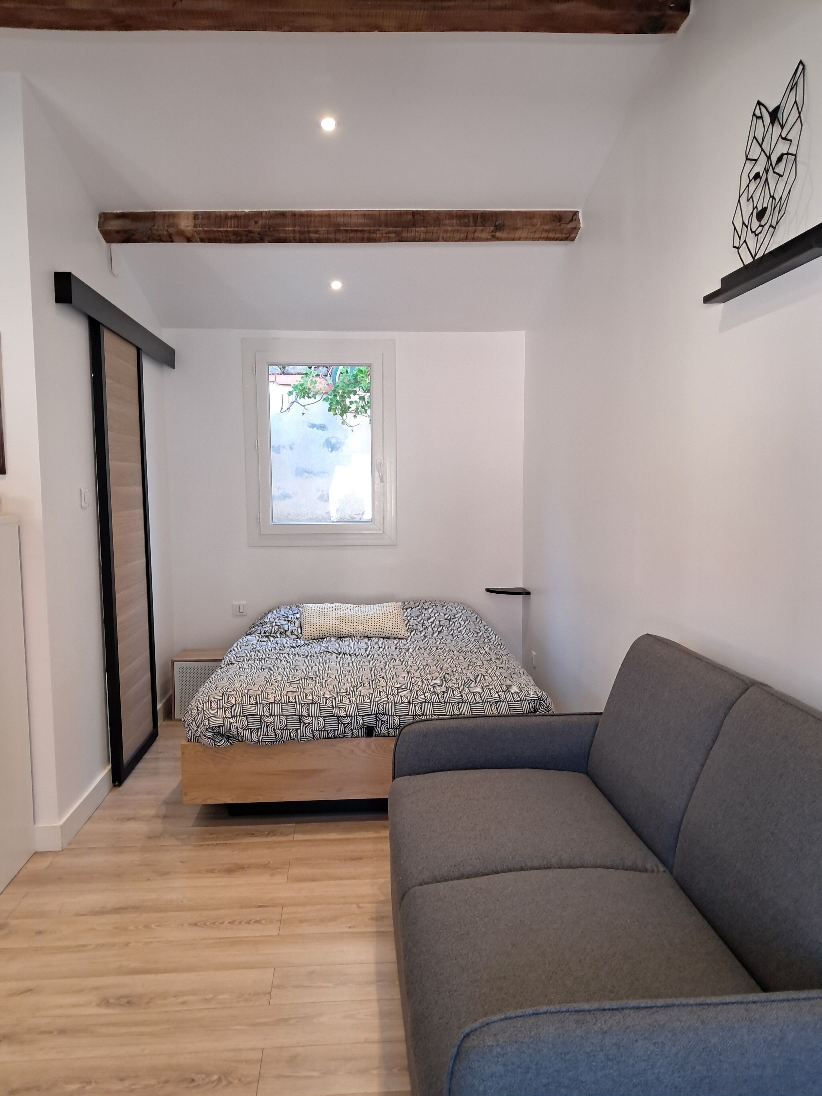 Een slaapkamer met een bed, een grijze bank, een raam met uitzicht op groen en een houten balk.
