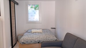 Ein Schlafzimmer mit einem Bett, einem grauen Sofa, einem Fenster mit Blick auf Grünflächen und einem Holzbalken an der Decke.