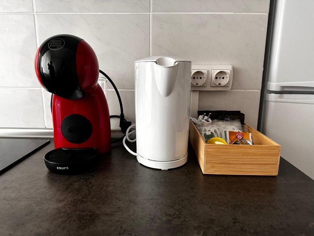 Una cocina con una cafetera roja y negra, un hervidor eléctrico blanco y una caja de madera con bolsitas de té y un limón.