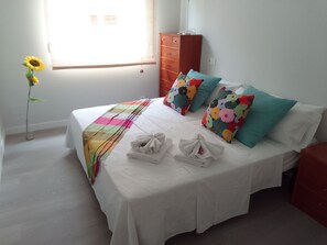 Un lit bien fait, agrémenté de coussins colorés, d’une couverture blanche et d’un vase à motifs floraux contenant un tournesol.