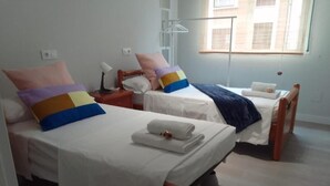 Habitación de hotel con dos camas, cada una con una colcha blanca y almohadas de colores. Hay una mesita de noche con una lámpara y una pequeña mesa con una taza. La habitación tiene una ventana con persianas y una lámpara colgante.