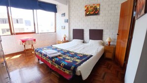 A hotel room with a large bed, a small table with a red tablecloth, two bedside lamps, a window with a blue curtain, and a white brick wall with a painting.