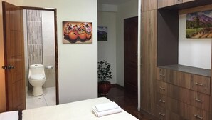 A hotel room with a bed, wooden wardrobe, a painting, a potted plant, and a toilet.