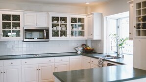 A kitchen with white cabinetry, a stainless steel microwave, and a window with a view of the outside. - Boothbay Harbor King Suite  - Room 13 at AQ Inn - Fireplace, Patio, Harbor View! (Boothbay Harbor)