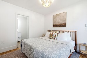 A bedroom with a bed, a bedside table, a nightstand, a wall-mounted artwork, and a door leading to another room. - Boothbay Harbor King Suite  - Room 13 at AQ Inn - Fireplace, Patio, Harbor View! (Boothbay Harbor)