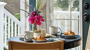 A wooden chair with a woven backrest, a round table with a white mug, a plate with a croissant, and a vase with flowers on a balcony. - Boothbay Harbor King Suite  - Room 13 at AQ Inn - Fireplace, Patio, Harbor View! (Boothbay Harbor)