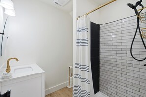 A bathroom with a white sink, gold faucet, and a shower with a black curtain. - Boothbay Harbor King Suite  - Room 13 at AQ Inn - Fireplace, Patio, Harbor View! (Boothbay Harbor)