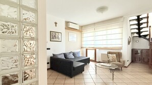 Ein Wohnzimmer mit einer Couch, einem Sessel und einem Couchtisch. Es gibt ein Fenster mit Jalousien, eine Wand-Klimaanlage und eine Treppe im Hintergrund.